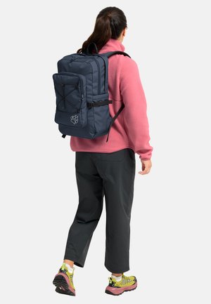Woman walking wearing a pink fleece jacket, black pants, yellow hiking shoes, and carrying a large navy blue backpack with a front pocket.