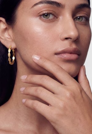 Ženska s svetlo poltjo in zelenimi očmi, ki nosi zlato obrobo, nežno dotika svojega obraza s čistimi, naravnimi nohti na enostavni ozadju.