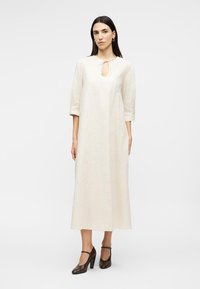 Γυναίκα που φορά ένα μακρύ κρεμ φόρεμα με μανίκια 3/4 και λαιμόκοψη με άνοιγμα, συνδυασμένο με σκούρα καφέ ψηλοτάκουνα παπούτσια Mary Jane, όρθια.