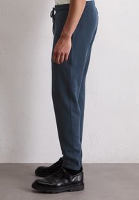 Blaue Sweatpants mit schlanker Passform, elastischem Bund und Kordelzugdetails. Mit Seitentaschen und gerippten Bündchen. Kombiniert mit schwarzen Schuhen.