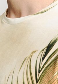 Beige T-shirt met lichte textuur, voorzien van groene plantillustraties. Ronde halslijn met een subtiele ribafwerking. Eenvoudig ontwerp zonder versieringen.