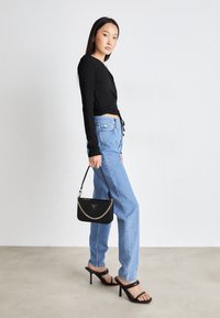 Haut noir à manches longues avec un détail noué, jean denim bleu clair, sac à main noir avec bandoulière chaîne, et sandales à talons noires. Textures lisses.