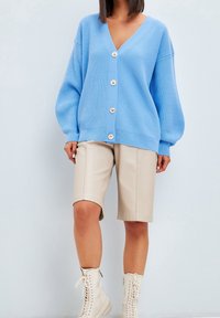 Jasnoniebieski prążkowany kardigan z dekoltem w serek, pięcioma dużymi złotymi guzikami oraz długimi bufkami. Połączony z beżowymi szortami i kremowymi botkami z wiązaniem.