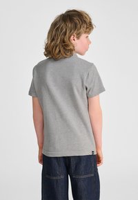 Szara koszulka z krótkim rękawem wykonana z bawełny, z okrągłym dekoltem i małą metką z logo przy dole. Nosi się ją z ciemnoniebieskimi dżinsami.