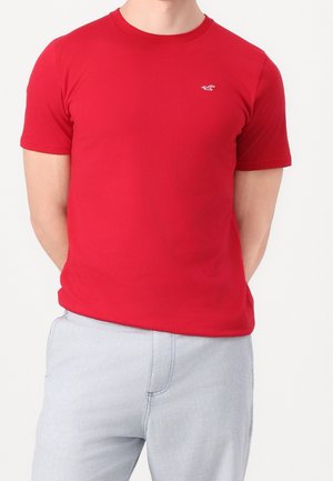 Homme portant un t-shirt rouge uni à manches courtes avec un petit logo blanc sur la poitrine et un pantalon gris clair, mains dans le dos.