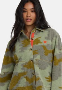 Donna con capelli lunghi e scuri che indossa una giacca in pile mimetica verde, grigia e senape con bottoni di legno e fodera rossa.