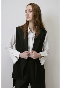 Svart ärmlös blazer med strukturerade slag över en vit skjorta. Skjortan har vida manschetter med guldknappaccent. Slät tygtextur.