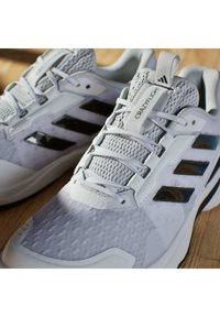 Białe buty sportowe z siateczkową cholewką, teksturowanym wzorem i trzema błyszczącymi czarnymi paskami. Uzupełnione o markową metkę i system sznurowania.