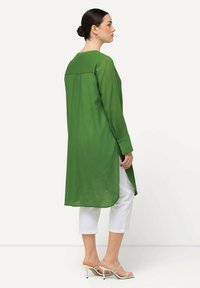 Green long-sleeve tunic made of lightweight fabric, featuring a straight cut, buttoned cuffs, and side slits, paired with white cropped pants.