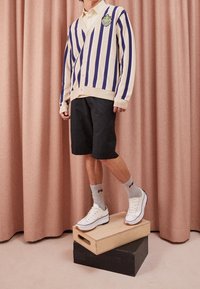 Gestreifter Cardigan in Creme und Lila mit V-Ausschnitt und Emblem, kombiniert mit schwarzen Shorts und weißen Sneakers sowie grauen Socken.