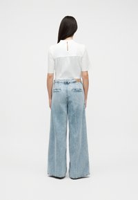 Γυναίκα με μακριά σκούρα μαλλιά που φοράει λευκό μπλουζάκι με κοντά μανίκια και ανοιχτόχρωμο τζιν παντελόνι με ψηλή μέση και φαρδιά μπατζάκια, στέκεται γυρισμένη με την πλάτη.