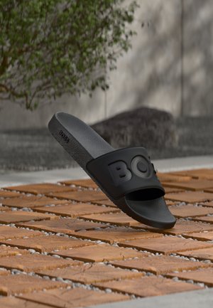 Melna slip-on sandale ar fakturētu zolīti un uzkrītošu "BOSS" logotipa siksniņu, novietota uz koka flīžu āra grīdas ar izplūdušu augu fonu.