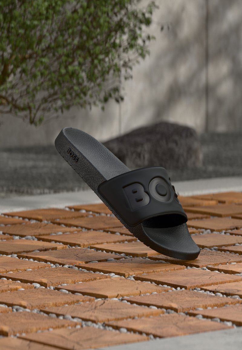Μαύρο σανδάλι τύπου slip-on με υφή στην σόλα και λουρί με έντονο λογότυπο "BOSS", τοποθετημένο σε ξύλινο πάτωμα με πλακάκια σε εξωτερικό χώρο και θολό φόντο με φυτά.