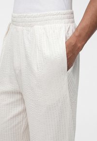 Κοντινό πλάνο υφασμάτινου λευκού παντελονιού με ελαστική μέση και πλαϊνή τσέπη, χέρι που ακουμπάει στην τσέπη με φόντο λευκό.