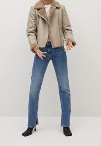 Donna con giacca in shearling beige, top bianco, jeans dritti blu con aperture alla caviglia, e stivali neri con tacco, in piedi contro uno sfondo neutro.