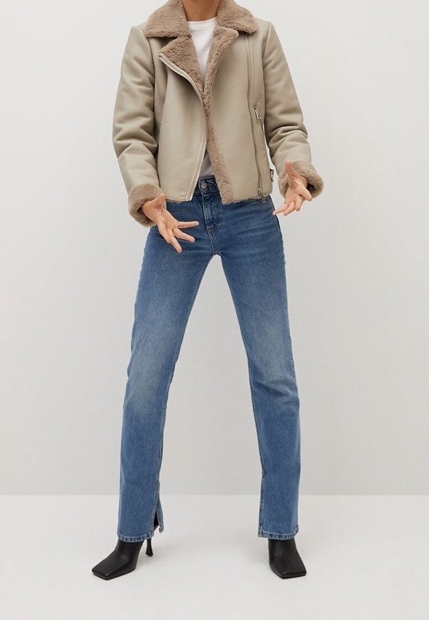 Donna con giacca in shearling beige, top bianco, jeans dritti blu con aperture alla caviglia, e stivali neri con tacco, in piedi contro uno sfondo neutro.