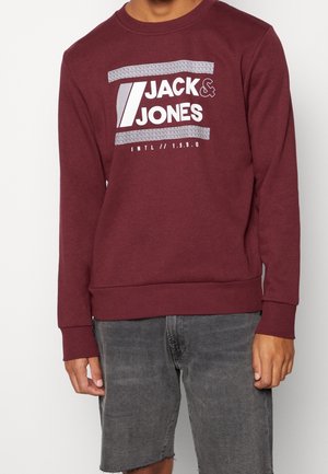 Person trägt ein kastanienbraunes Sweatshirt mit dem weißen Aufdruck "Jack & Jones Intl 1990" und eine graue Jeansshorts.