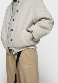 Sweatshirt gris avec fermeture à boutons et poches latérales, associé à un pantalon de survêtement beige avec un détail de logo brodé et une taille ajustable.
