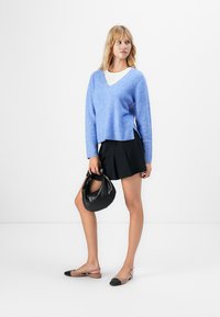 Žena u plavom V-izrezu puloveru, crnoj plisiranoj suknji, dvobojnim slingback balerinkama, drži crnu kožnu torbu dok stoji protiv bijele pozadine.
