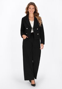 Giacca nera sartoriale con chiusura a doppio petto con bottoni argentati, abbinata a pantaloni neri a gamba larga. Tessuto morbido e design strutturato.