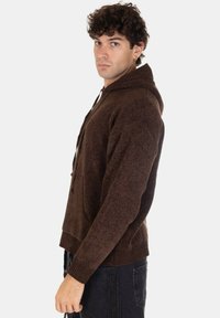 Felpa con cappuccio marrone realizzata in un tessuto a maglia testurizzato, dotata di una tasca frontale e polsini a coste. Mostrata su un modello maschile di profilo.