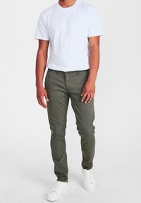 Man står klädd i en enkel vit t-shirt, olivgröna byxor och vita sneakers mot en vit bakgrund.