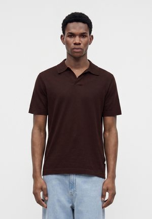 Jeune homme debout face à l’avant portant un polo à manches courtes marron foncé et un jean bleu clair sur un fond uni.