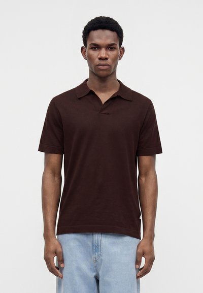 Jeune homme debout face à l’avant portant un polo à manches courtes marron foncé et un jean bleu clair sur un fond uni.