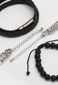 Quatre bracelets : un bracelet en cuir noir tressé, une chaîne en argent, un bracelet en argent à maillons avec un fermoir en forme de homard, et un bracelet extensible en perles noires.