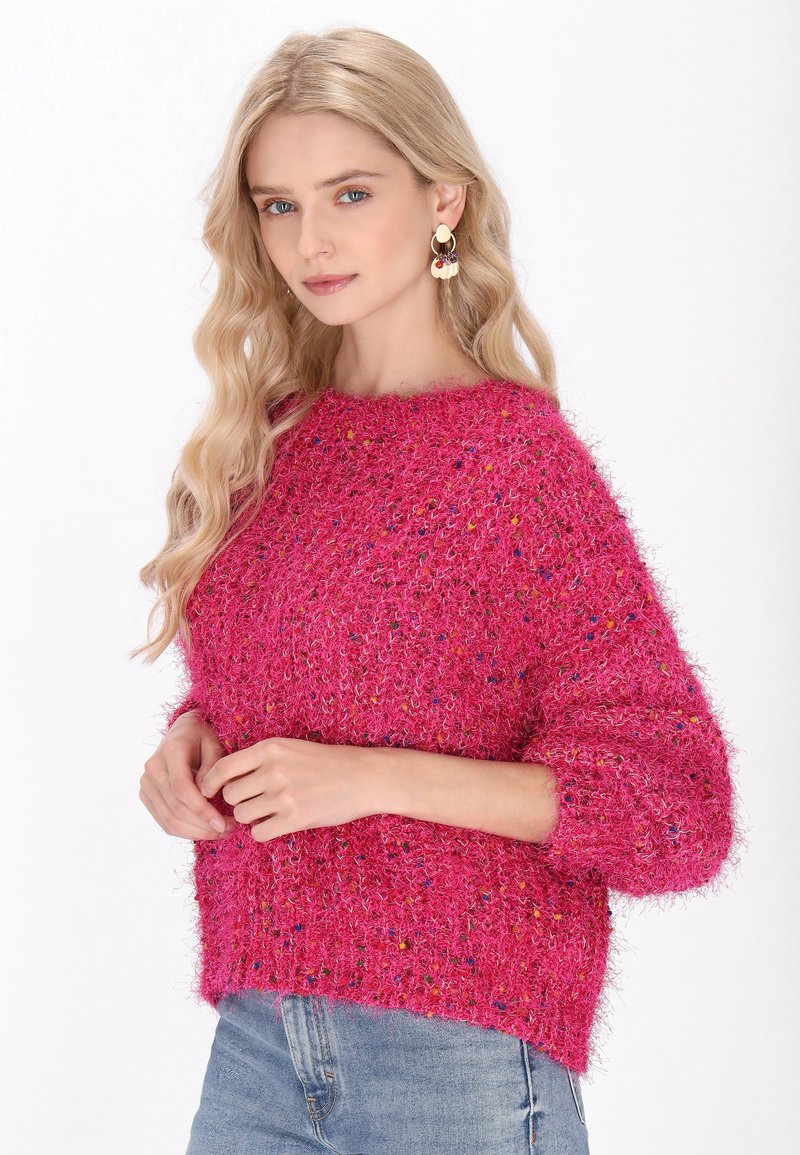 Puszysty różowy sweter o luźnym kroju i szerokich rękawach, z kolorowymi nitkami rozrzuconymi w całej strukturze. W połączeniu z jasnoniebieskimi dżinsami.