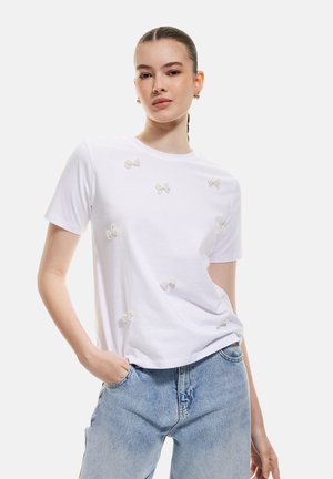 White cotton T-shirt with short sleeves featuring embroidered white bows scattered across the front. Paired with light blue denim jeans.