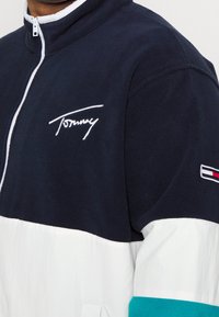 Granatowo-biała kurtka polarowa z wysokim kołnierzem, z białym haftowanym logo oraz konstrukcją typu color-block z turkusowymi i czerwonymi akcentami na rękawach.