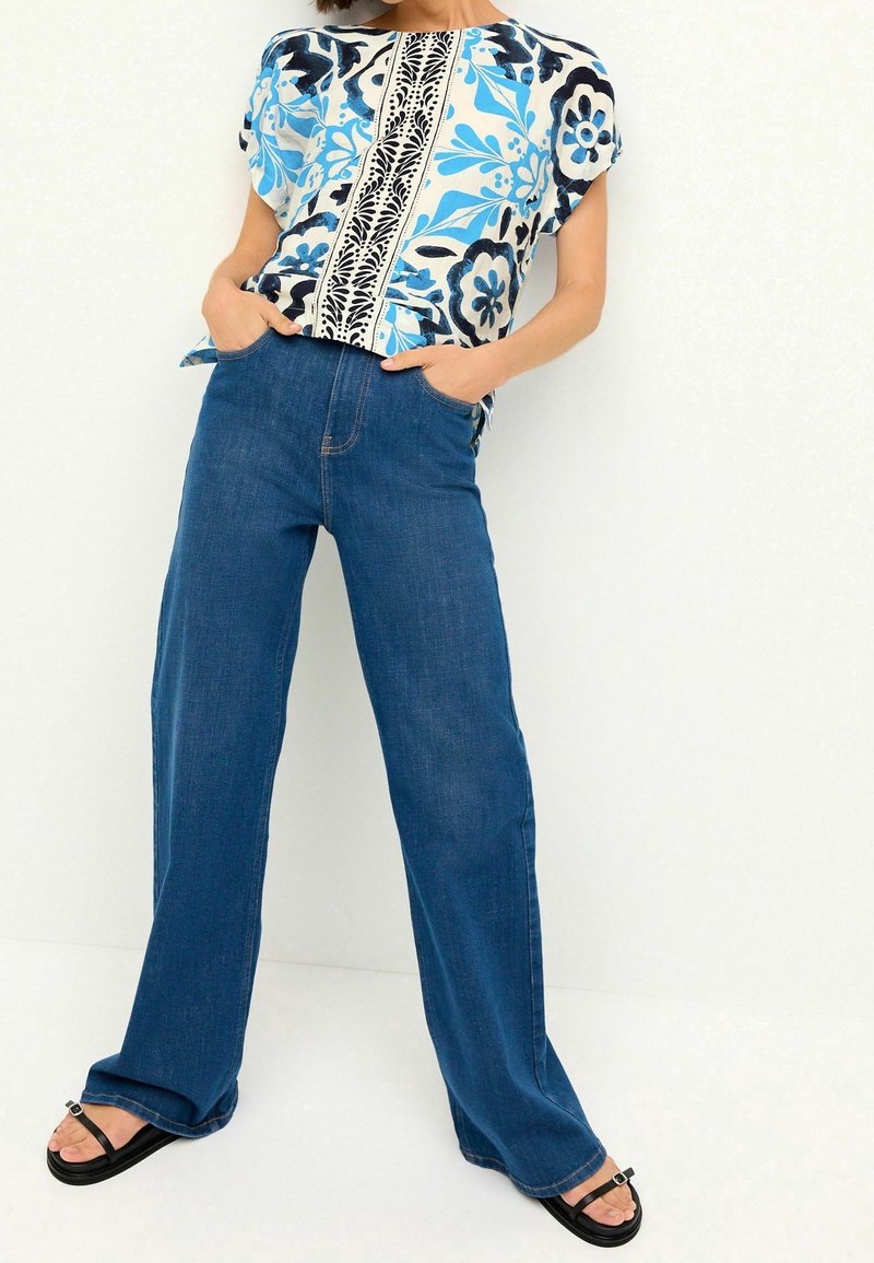 Blusa de manga corta en un patrón floral azul y blanco, combinada con pantalones de mezclilla azul de pierna ancha y alta cintura, y sandalias planas negras.