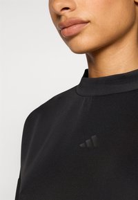 Crni atletski top s visokim izrezom, mekanom teksturom tkanine i suptilnim crnim Adidas logom na prsima.