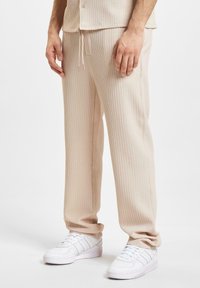Beige ribbede bukser med snøre i taljen, tapered pasform og sidelommer, parret med hvide sneakers. Teksturert stof er synligt.