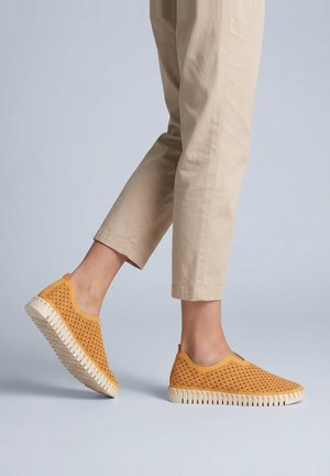 Person trägt beige Hose und senfgelbe Slip-On-Schuhe mit weißer Sohle und Wellenmuster, geht auf einem hellgrauen Boden.