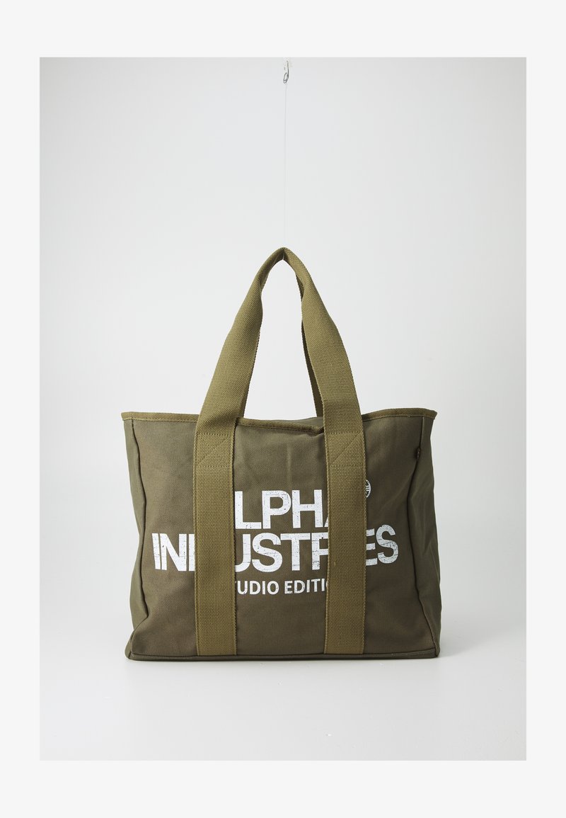 Oliivivihreä kankaasta valmistettu kassi, jossa on kaksi tukevaa kantohihnaa. Kassin etupuolella on valkoisella painettu teksti "ALPHA INDUSTRIES STUDIO EDITION." Lohkoperäinen muoto ja teksturoitu pinta.