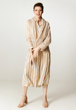 Kobieta stojąca i uśmiechająca się, ubrana w beżową sukienkę w paski, jasnobrązowy otwarty sweter z dzianiny oraz jasnobrązowe sandały na obcasie na jednolitym tle.