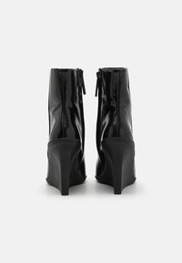 Une paire de bottines brillantes à talons hauts noires avec des fermetures éclair sur les côtés intérieurs, présentées de dos sur un fond blanc uni.