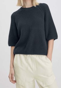 Femme portant un pull noir à manches courtes en maille texturée et un pantalon beige clair à cordon de serrage avec de grandes poches, debout devant un fond uni.