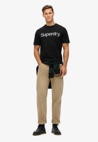 Camisa de algodón negra con el logo "Superdry", pantalones de sarga beige, camisa de cuadros verde atada a la cintura y botas de cuero negras. Diseño minimalista.