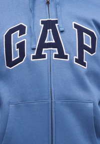 Sweat à capuche zippé bleu avec de grandes lettres "GAP" bleu marine et blanches délimitées sur la poitrine et des poches kangourou à l'avant.