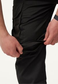Schwarze Hosen aus strukturiertem Stoff, mit einer seitlichen Reißverschlusstasche, die von einer Hand geöffnet wird. Das Design zeichnet sich durch einen körpernahen Schnitt aus.