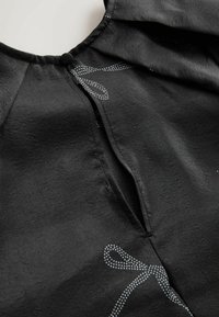 Juodas audinys su šiek tiek blizgesio, turintis apykaklę su lenkimu ir dekoratyviu balto taškuoto kaspino raštu. Matomas pamušalo siūlė.