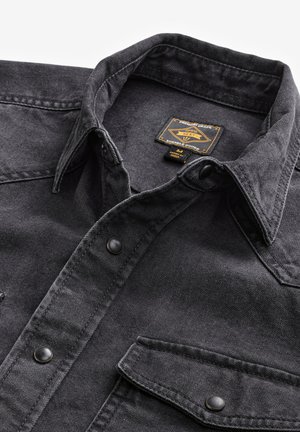 Svart denimskjorta med spetsig krage, försedda med två bröstfickor och svarta tryckknappar. Texturen verkar robust med en slät yta.