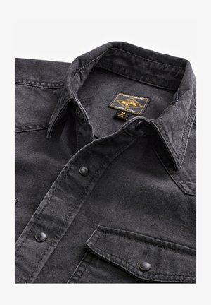 Chemise en denim noire avec un col pointu, comportant deux poches poitrine et des boutons-pression noirs. La texture semble robuste avec une finition lisse.