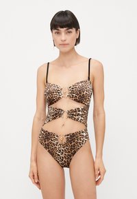 Donna che indossa un costume da bagno a fascia con stampa leopardata, dettagli in oro e sottili spalline nere, in piedi su uno sfondo semplice.
