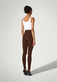 Bruna högmidjade leggings med en slät yta, matchade med en vit tanktopp och svarta högklackade skor, sedda bakifrån.