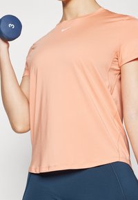 Camiseta deportiva de manga corta color melocotón claro con cuello redondo y un sutil logo de Nike. La persona sostiene una pesa azul con la etiqueta "3".