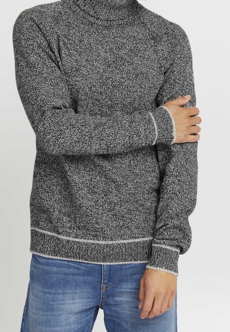 Homme portant un pull en laine gris avec bordure blanche, tenant son bras gauche avec sa main droite, vêtu d'un jean bleu sur un fond uni.